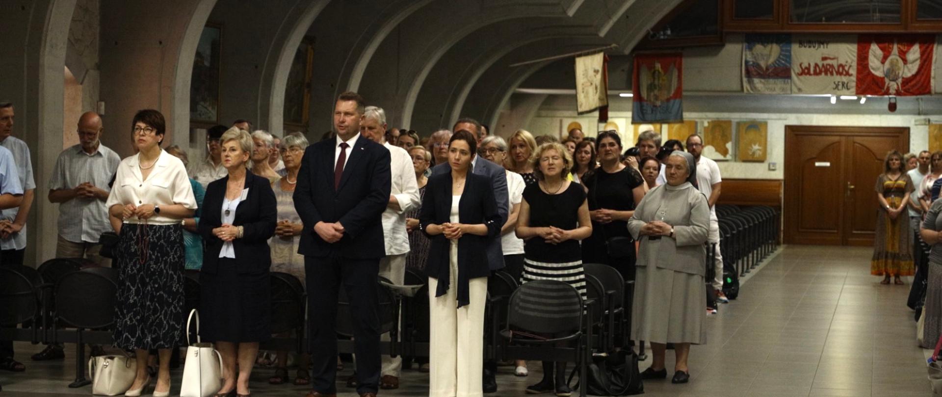 Minister i inni uczestnicy pielgrzymki stoją pod żebrowanym sufitem.