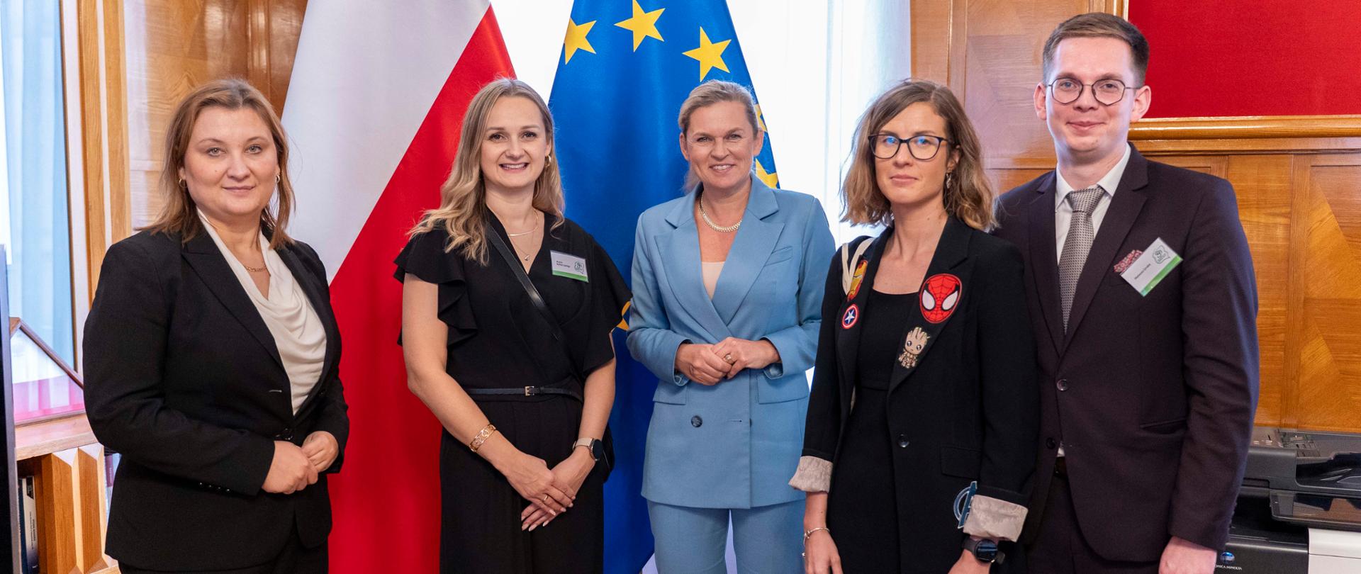 Pięć osób, w tym cztery kobiety i jeden mężczyzna, stoi w rzędzie i pozuje do zdjęcia w pomieszczeniu z drewnianymi panelami. Za nimi widoczne są flagi Polski i Unii Europejskiej. Jedna z kobiet ma na marynarce przypinki z postaciami z komiksów.