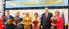 Minister Czarnek w otoczeniu grupy kolorowo ubranych osób stoi pod ścianą z napisem Witamy w nowym przedszkolu.