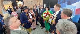 Minister i ubrana na zielono kobieta stoją i pokazują bukiet kwiatów i laleczki w ludowych strojach, przed nimi duża grupa ludzi, obok ceglana ściana.