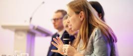 Minister funduszy i polityki regionalnej Katarzyna Pełczyńska-Nałęcz siedzi przy stole i przemawia do mikrofonu trzymanego w dłoniach