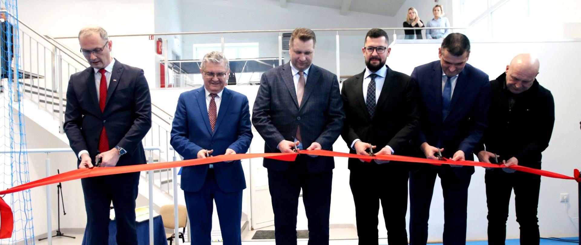 Minister stoi w grupie 5 mężczyzn, wszyscy mają nożyczki i razem przecinają czerwoną wstęgę, za nimi w tle biała klatka schodowa.