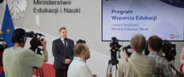 Minister stoi i mówi do mikrofonu, za nim ekran z napisem Program wsparcia edukacji, przed nim dziennikarze z kamerami.
