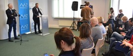 Wiceminister zdrowia Janusz Cieszyński odpowiada na pytania dziennikarzy podczas konferencji prasowej w Centrum Systemów Informacyjnych Ochrony Zdrowia. Na zdjęciu wiceminister za mównicą, na tle roll-upu Ministerstwa Zdrowia. Na przeciwko niego dwa rzędy dziennikarzy oraz kamery.