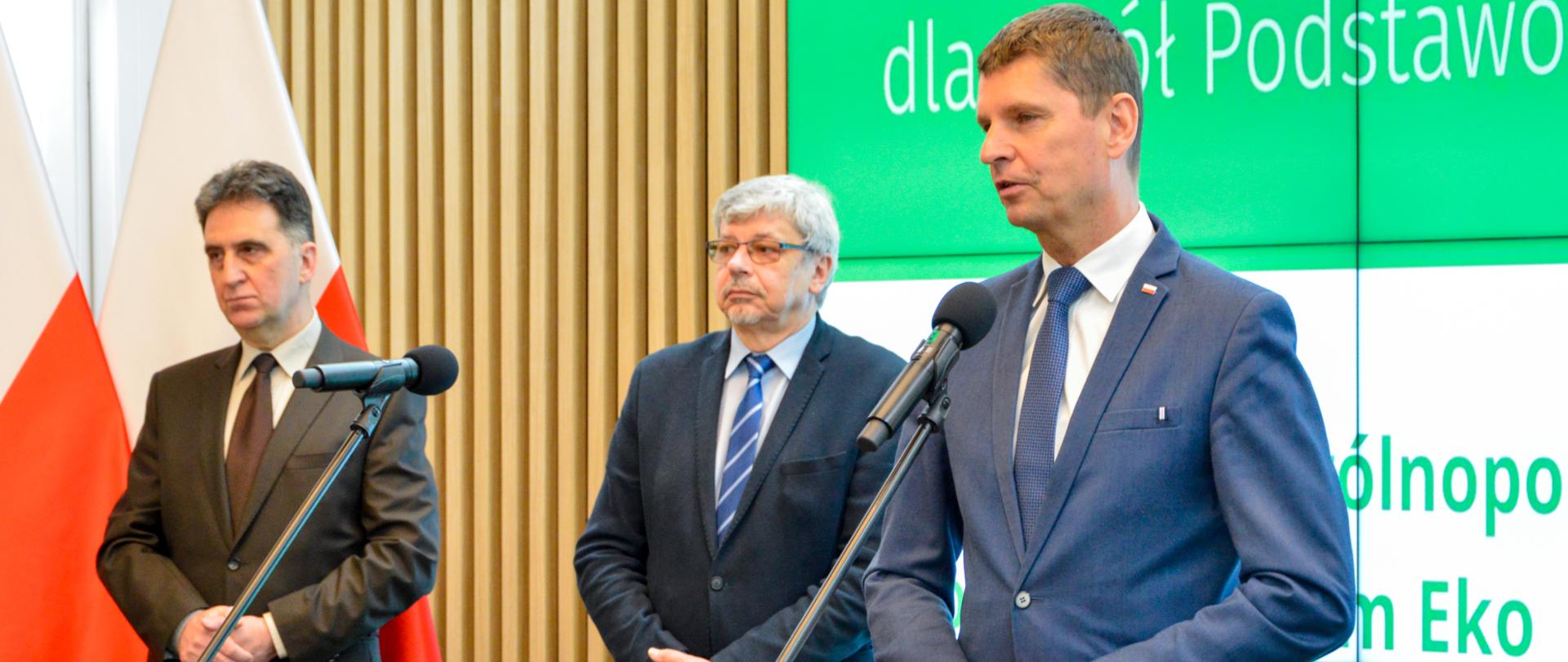 Trzej mężczyźni ubrani w garnitury i stojący przy mikrofonach. W tle z lewej strony biało-czerwona flaga, boazeria i duży biało-zieony ekran. 