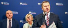 Minister Czarnek stoi i mówi do mikrofonu, za nim stoi wiceminister Machałek w białym ubraniu i mężczyzna w szarym garniturze, za nimi granatowa ściana.