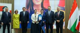 Pod ścianą z pionowych drewnianych deseczek, na której wisi wielki ekran na którym widać twarz kobiety, stoi grupa siedmiu osób - pięciu mężczyzn w garniturach i dwie kobiety, obok nich węgierska flaga.