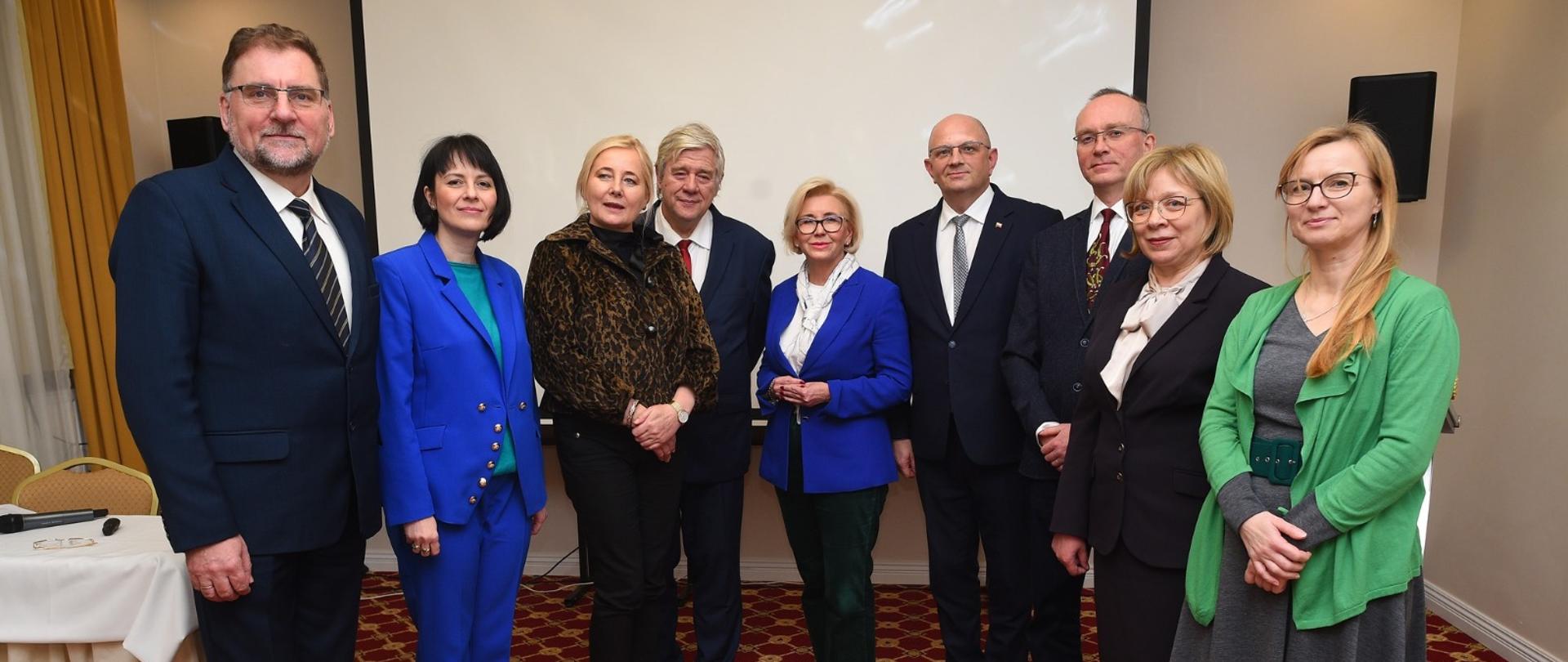 Kilka osób, kolorowo ubrane kobiety i mężczyźni w garniturach, stoi w pokoju z jasnymi ścianami i podłogą w czerwone kwadraty, z lewej nakryty stół.