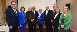 Kilka osób, kolorowo ubrane kobiety i mężczyźni w garniturach, stoi w pokoju z jasnymi ścianami i podłogą w czerwone kwadraty, z lewej nakryty stół.