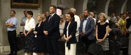 Minister stoi razem z innymi uczestnikami pielgrzymki, za nimi kamienne ściany na których wiszą obrazy.