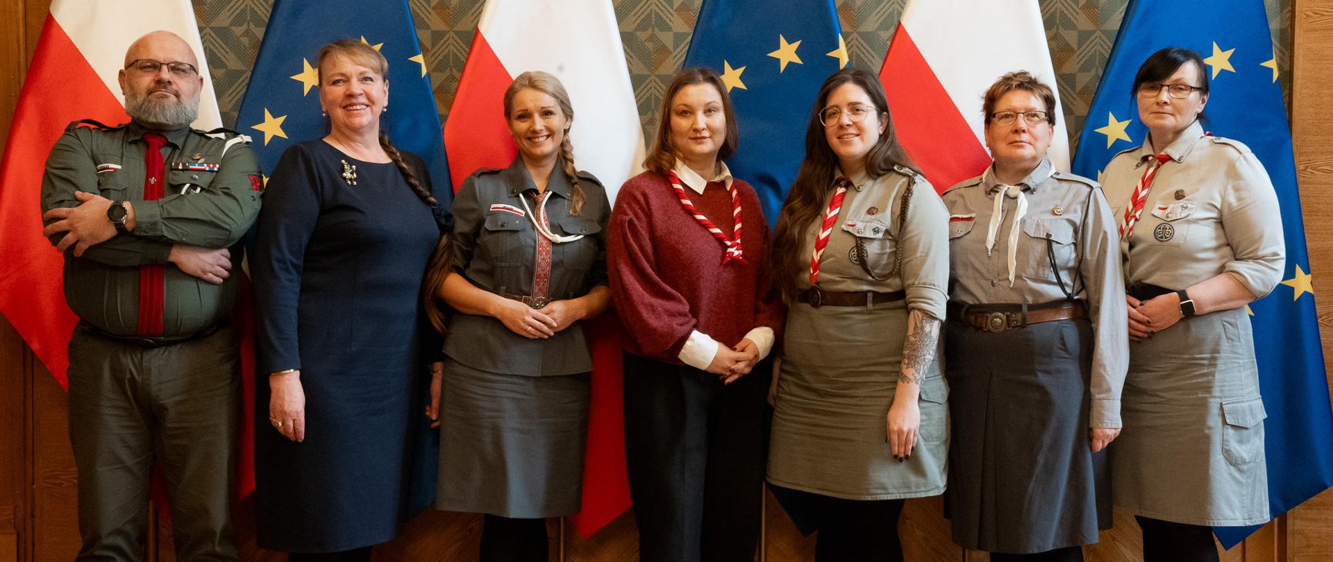 Grupa kobiet i mężczyzn w mundurach harcerskich pozuje do zdjęcia. w tle flagi Polski i UE