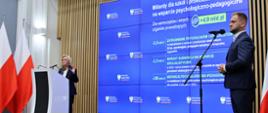 Widok z boku pod kątem na salę, wiceminister Machałek stoi za mównicą i mówi do dwóch mikrofonów, za nią wielki niebieski ekran z napisem Miliardy dla szkół i przedszkoli, z boku przed mikrofonem na stojaku stoi mężczyzna w garniturze.