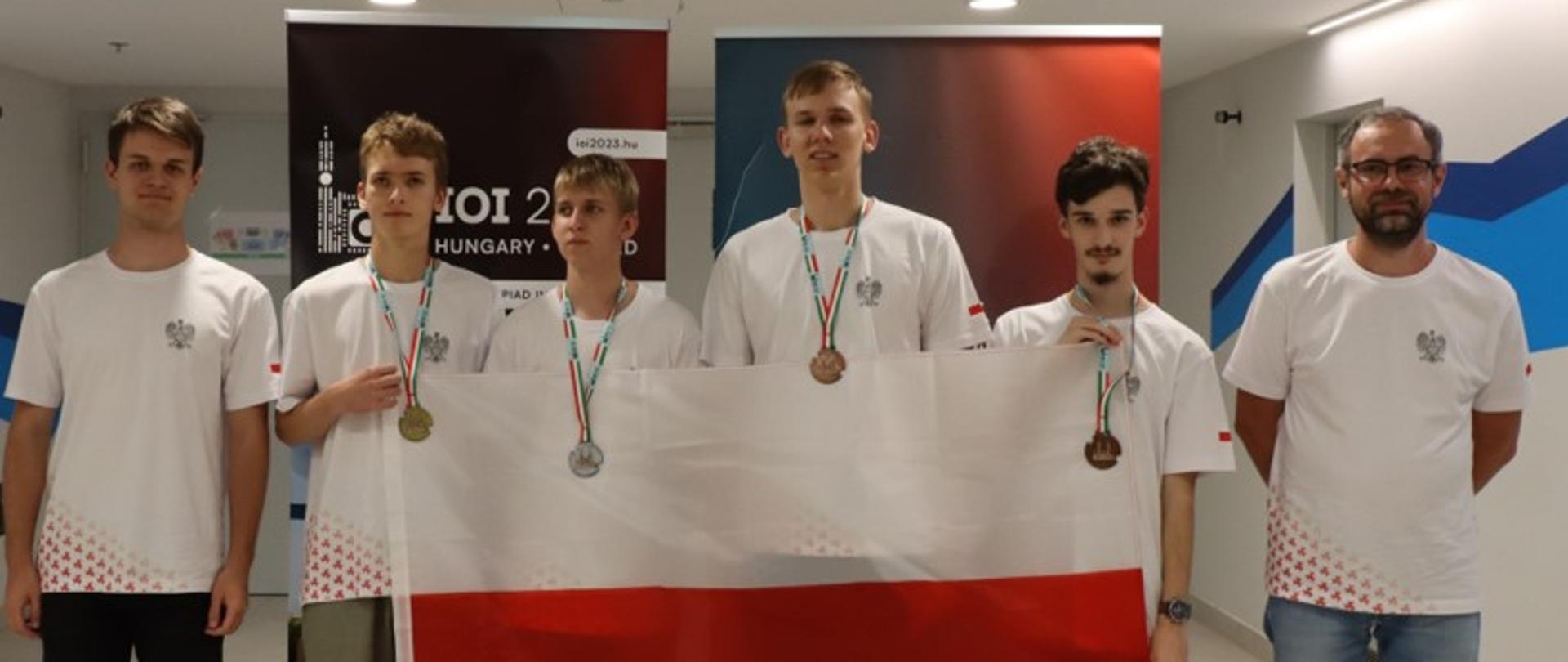 Sześciu młodych ludzi w białych koszulkach stoi przed kolorowym banerem, trzymają polską flagę.
