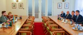 Widok na salę, po obu stronach drewnianego stołu siedzą grupy ludzi, przed grupa po lewej soi mała ukraińska flaga, po prawej - polska, za stołem duże przeszklone drzwi.