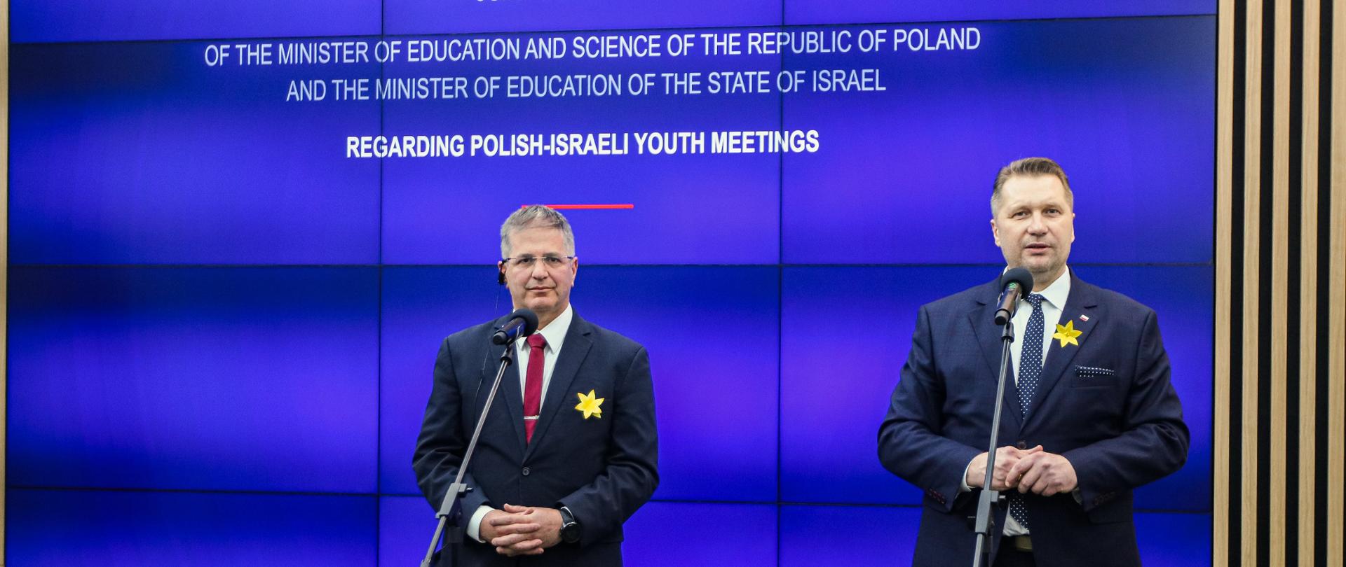 Minister Czarnek i mężczyzna w czarnym garniturze stoją przy mikrofonach na stojakach, obaj mają przypięte do garniturów żółte papierowe kwiaty, za nimi wielki telebim z napisem Joint declaration na niebieskim tle.