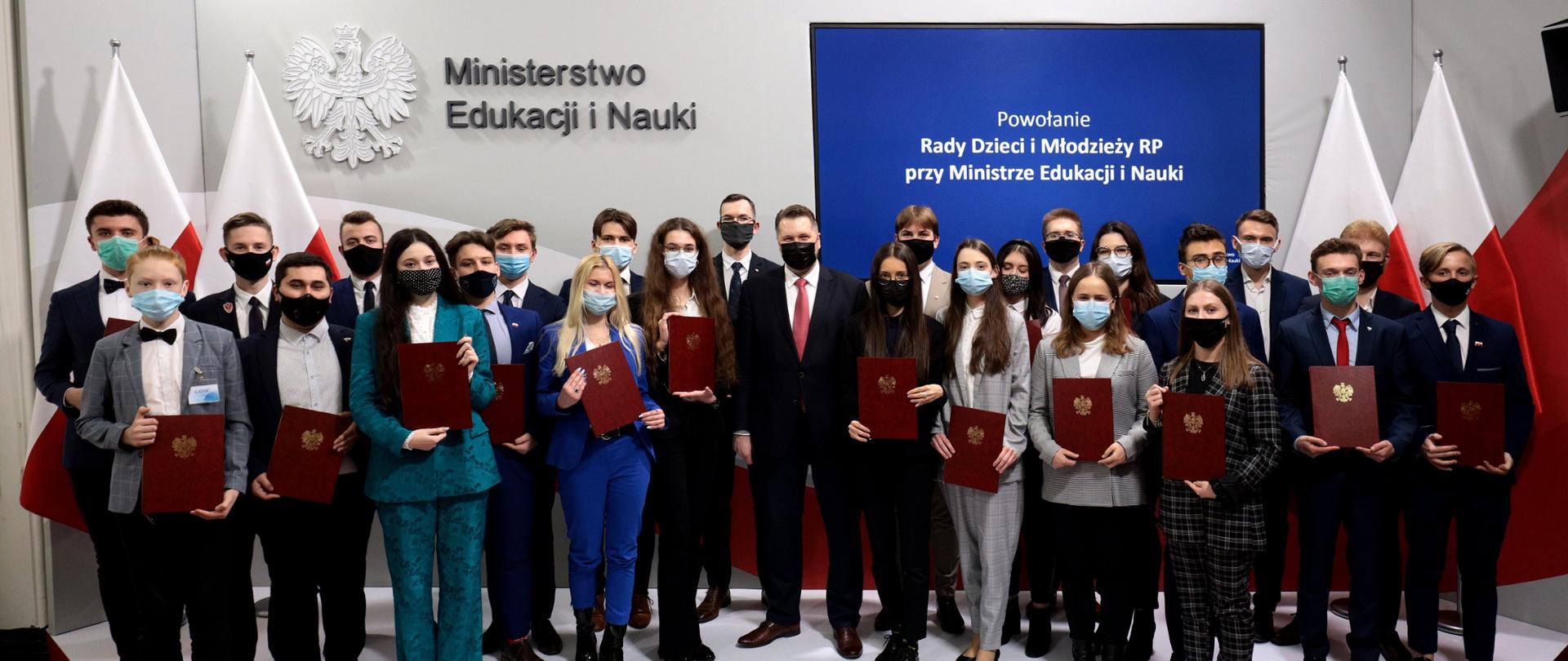 Na podwyższeniu stoi minister Czarnek otoczony dużą grupą młodych elegancko ubranych ludzi. W ręku mają trzymają oni czerwone teczki ze złotymi orłami. Wszyscy mają na twarzach maseczki. W tle biało-czerwone flagi, orzeł, napis Ministerstwo Edukacji i Nauki oraz monitor z grantowym tłem i napisem Powołanie Rady Dzieci i Młodzieży RP przy Ministrze Edukacji i Nauki. 