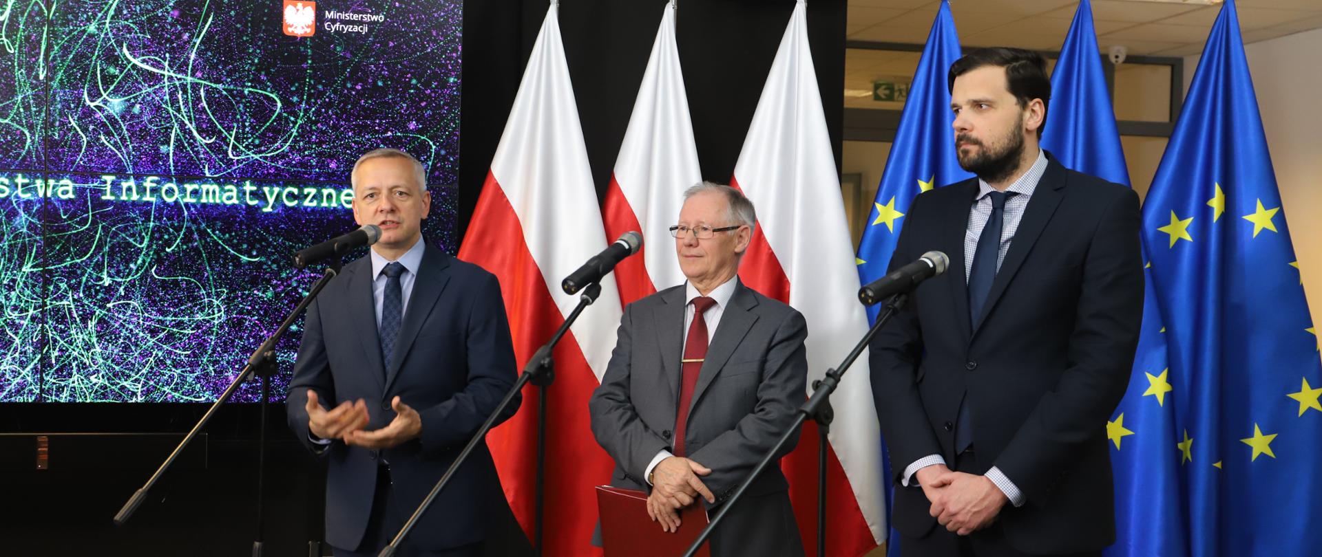 Od lewej stoją przed mikrofonami: minister cyfryzacji, Marek Zagórki, dyrektor Centrum Projektów Polska Cyfrowa, Wojciech Szajnar oraz rektor Politechniki Łódzkiej, prof. Sławomir Wiak. W tle flagi Polski i UE.