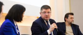 Widok z boku, przy stolikach siedzi trzech mężczyzn i kobieta ubrana na niebiesko, jeden z nich - wiceminister Rzymkowski mówi do trzymanego w ręku bezprzewodowego mikrofonu.