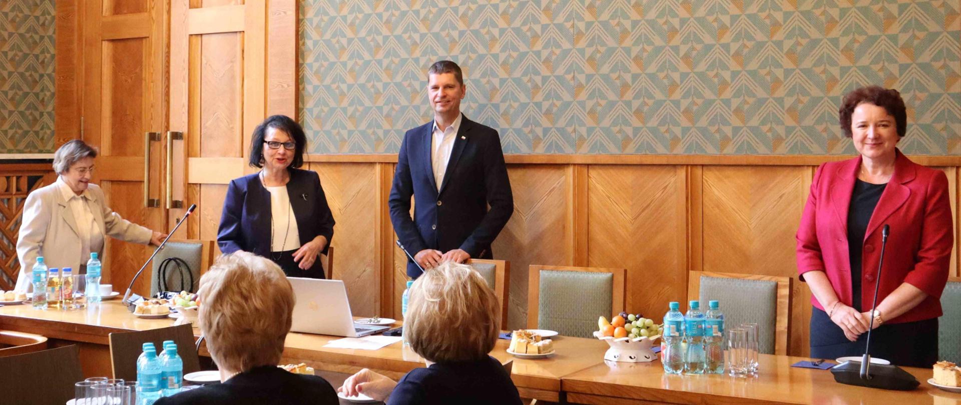 Duża sala ze ścianami w zielony wzorek, przy drewnianych stołach siedzi dużo elegancko ubranych osób. Na stole filiżanki, talerzyki i butelki z wodą. U szczytu stołu stoi wiceminister Piontkowski i dyrektor Lizut