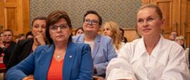 Widok z przodu na dużą salę, na ustawionych w rzędy krzesłach siedzą ludzie. W pierwszym rzędzie minister Barbara Nowacka i minister Izabela Leszczyna. Za nimi wiceminister Katarzyna Lubnauer i Paulina Piechna-Więckiewicz