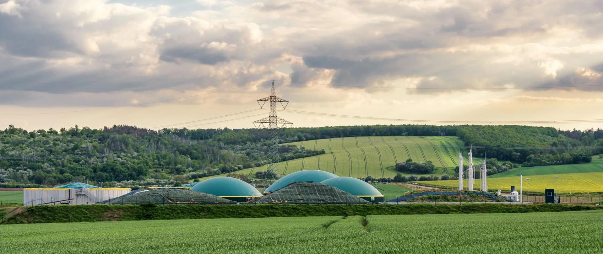Instalacja biogazowa z charakterystycznymi kopułami fermentacyjnymi położona w otoczeniu pól uprawnych. W tle widoczne są lasy oraz infrastruktura elektroenergetyczna.