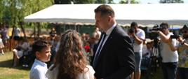 Minister rozmawia z dwójką dzieci, stoją na trawie, za nimi namiotowy daszek.