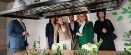 Minister funduszy i polityki regionalnej Katarzyna Pełczyńska-Nałęcz stoi przed stołem z kwiatami, obok niej stoją osoby, z którymi zwiedza Branżowe Centrum Umiejętności (BCU) Przemysłu Drzewnego w Kaliszu

