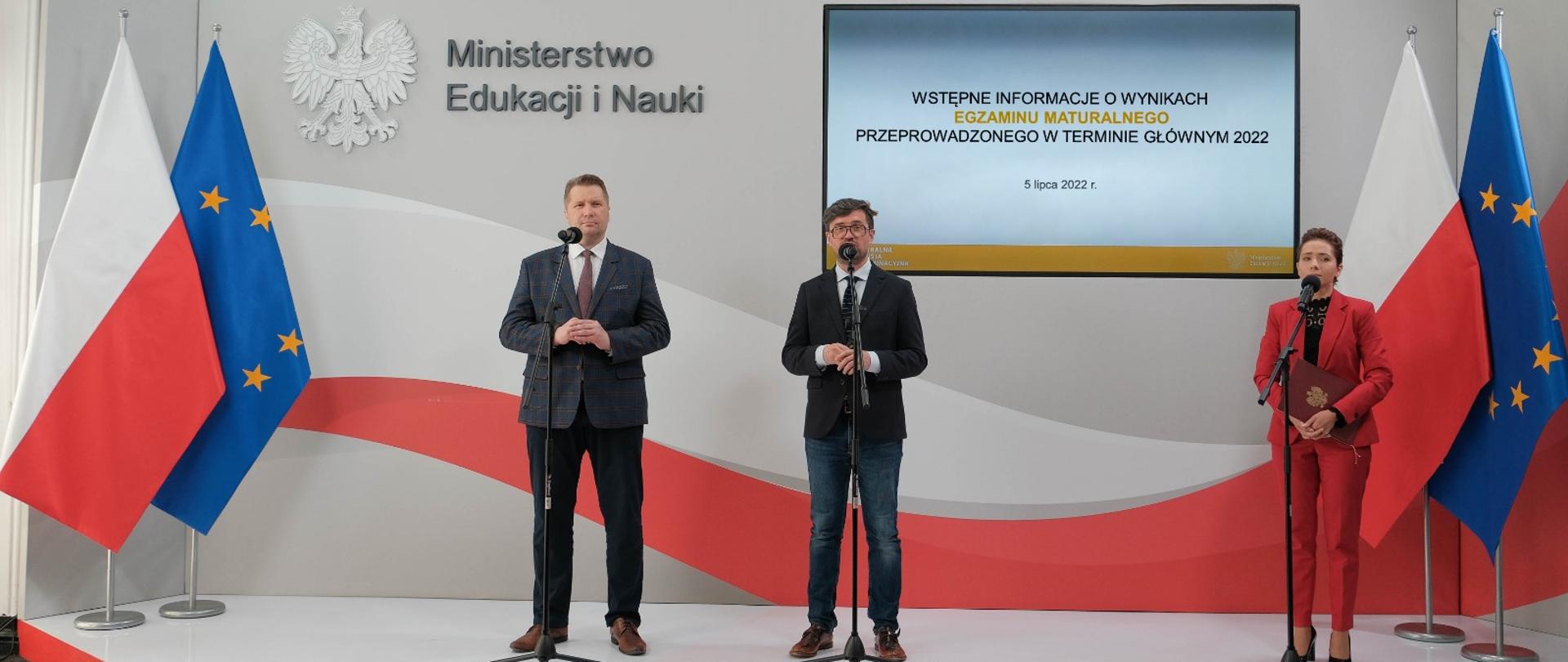Widok z przodu - na podwyższeniu stoją minister Czarnek, mężczyzna w czarnym garniturze i kobieta ubrana na czerwono, za nimi flaga Polski.