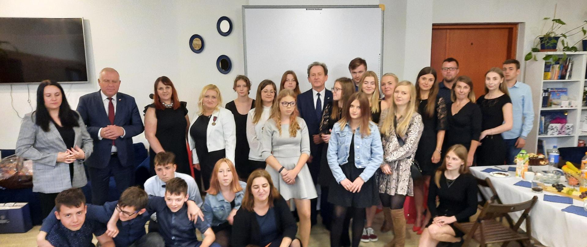 Sala lekcyjna. Wiceminister Bernacki w otoczeniu grupy dzieci i nauczycieli. Część z nich stoi i siedzi, część kuca. Grupa jest ubrana w uroczyste stroje. 