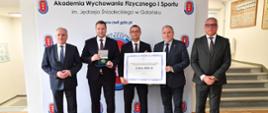 Uczestnicy wizyty stoją przy schodach, jeden trzyma symboliczny czek.