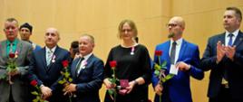 Pod żółtą ścianą stoi szereg kilku osób trzymających w rękach po jednej czerwonej róży, obok stoi minister Czarnek i bije brawo.