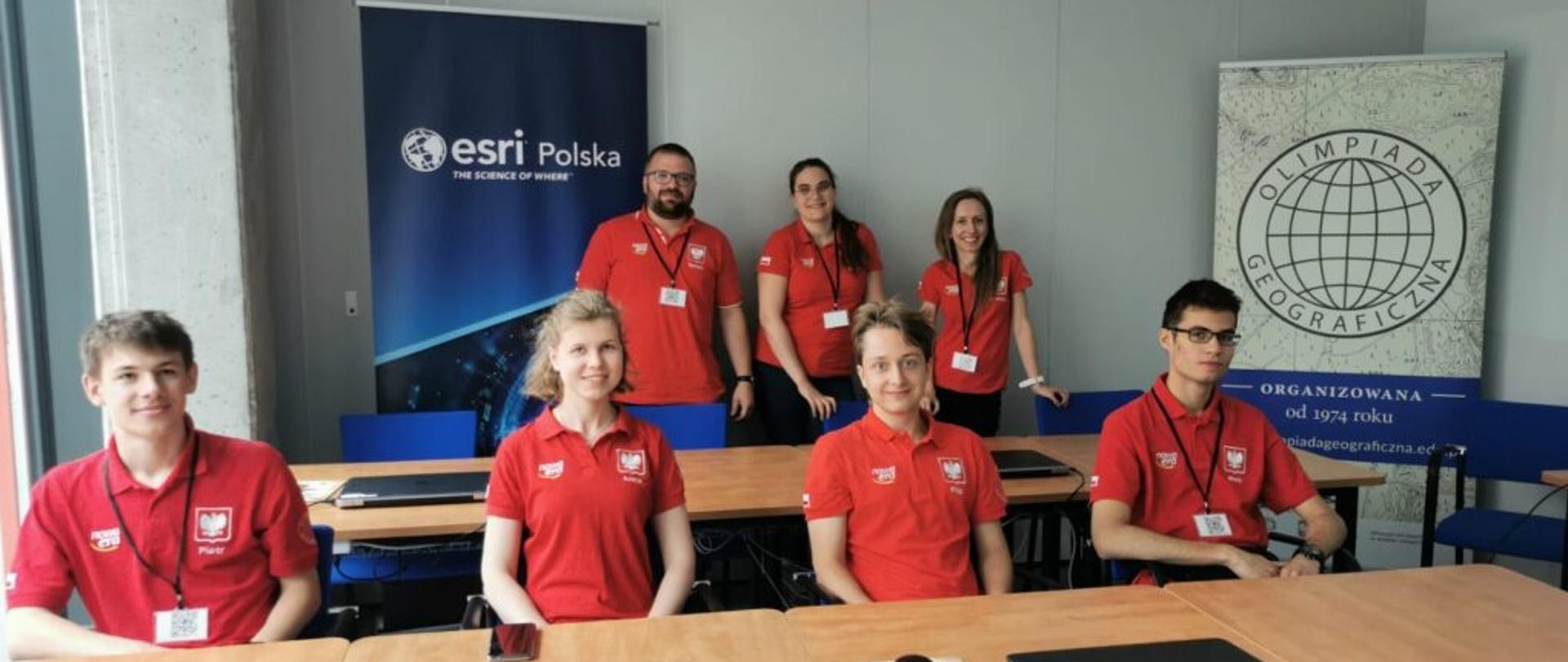 Grupa młodych ludzi w czerwonych koszulkach z godłem Polski siedzi przy ławkach, za nimi plakaty z napisami ESRI Polska i Olimpiada geograficzna.