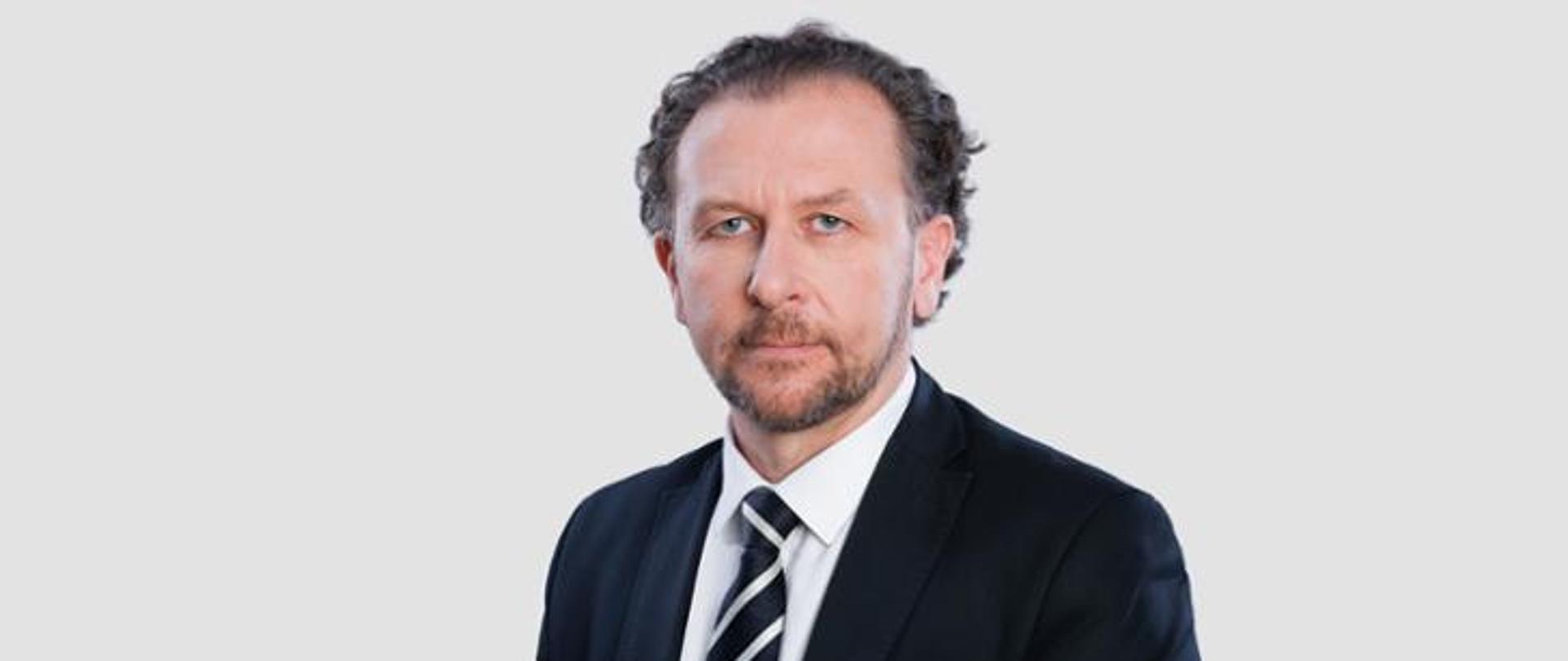 Na tle jasnoszarej ściany stoi dyrektor Bartold w czarnym garniturze.