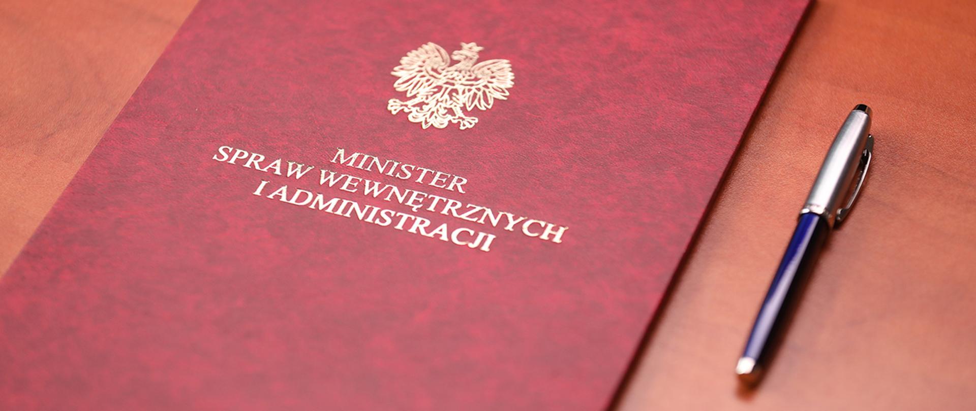 Teczka MSWiA z motywem orła z Godła RP oraz podpisem "Minister spraw wewnętrznych i administracji", obok leży długopis