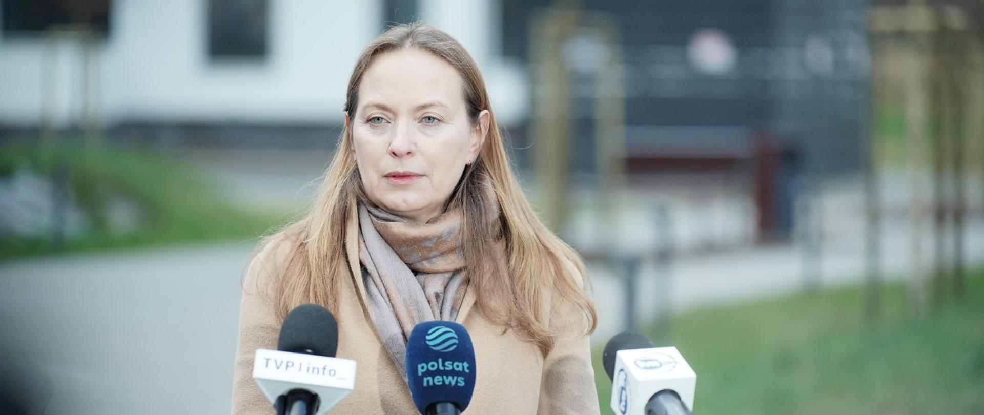 Na zdjęciu minister Katarzyna Pełczyńska-Nałęcz stoi przed mikrofonami i odpowiada na pytania dziennikarzy