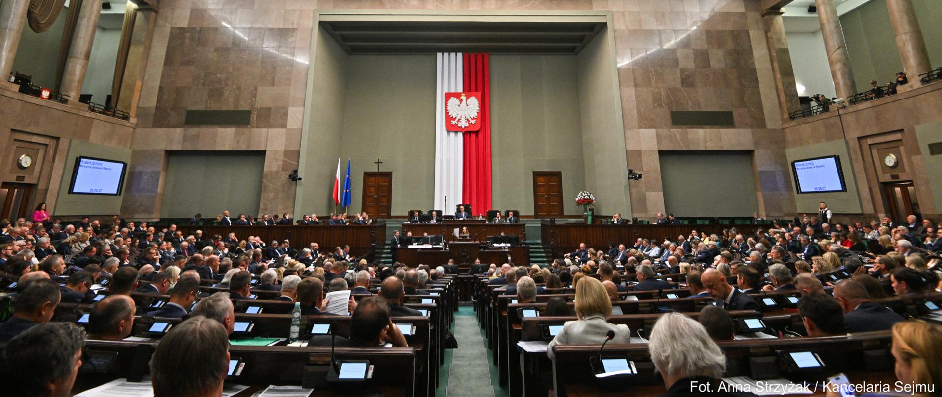 66. posiedzenie Sejmu. Zdjęcie przedstawia widok na salę plenarną z polską flagą w tle. Na sali, w ławach siedzą posłowie, przedstawiciele rządu, marszałek i wicemarszałkowie. 