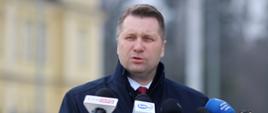 Minister Czarnek stoi i mówi do kilku mikrofonów, za nim w tle zarys budynku stacji kolejowej.