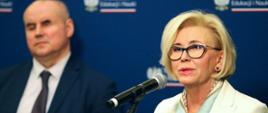 Wiceminister Machałek w białym ubraniu stoi i mówi do mikrofonu, z lewej strony stoi mężczyzna w garniturze, za nimi granatowa ściana.
